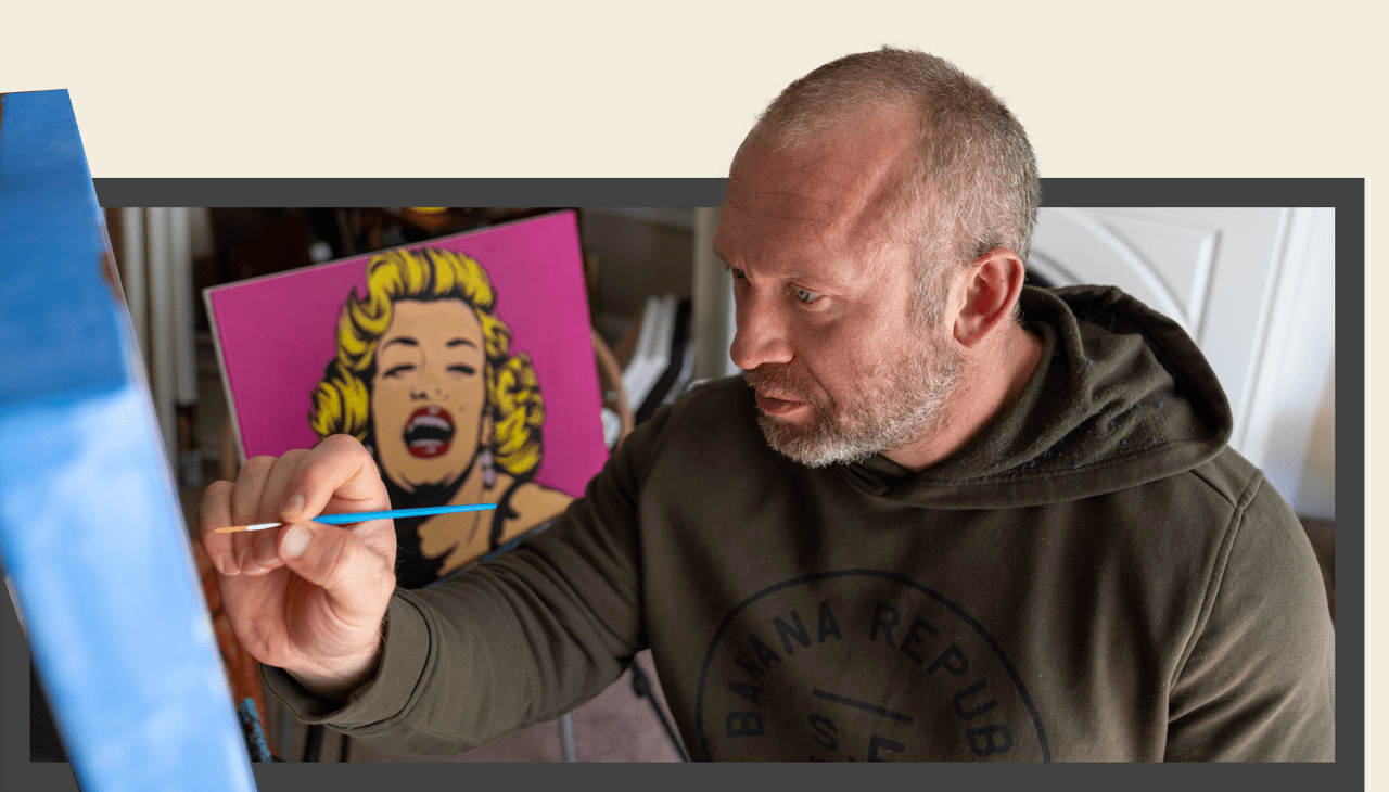 Artist Andrew French painting in his studio, focused on a canvas while a colorful pop art portrait of Marilyn Monroe appears in the background. The image captures his creative process and signature pop-inspired artistic style.