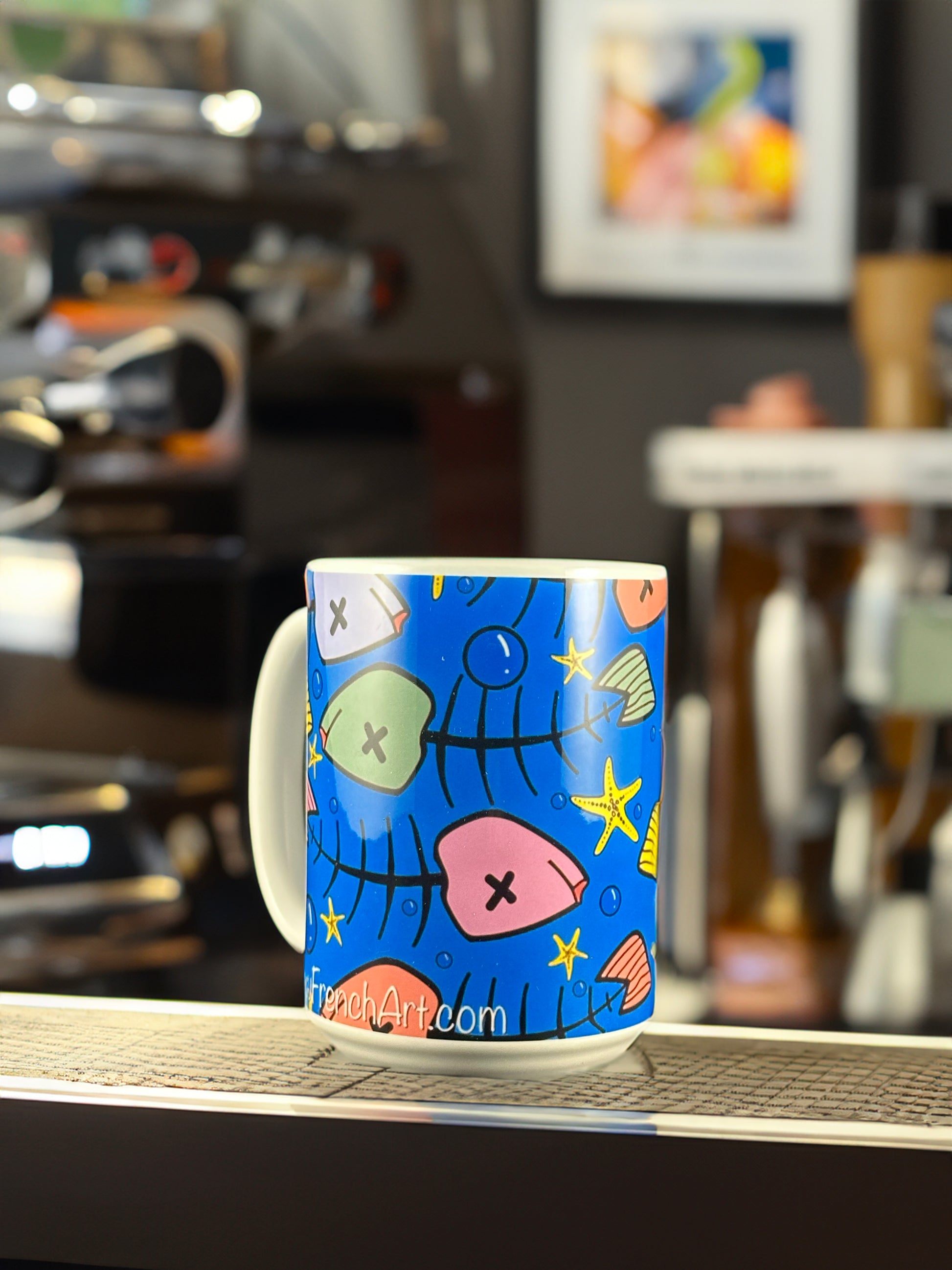 A bold ceramic mug featuring Andrew French’s vibrant “Toxic Blue Xmas Fish” design. The artwork displays whimsical fish bones, seashells, and starfish in bright holiday colors against a deep ocean-blue background — a playful fusion of coastal art and festive cheer.