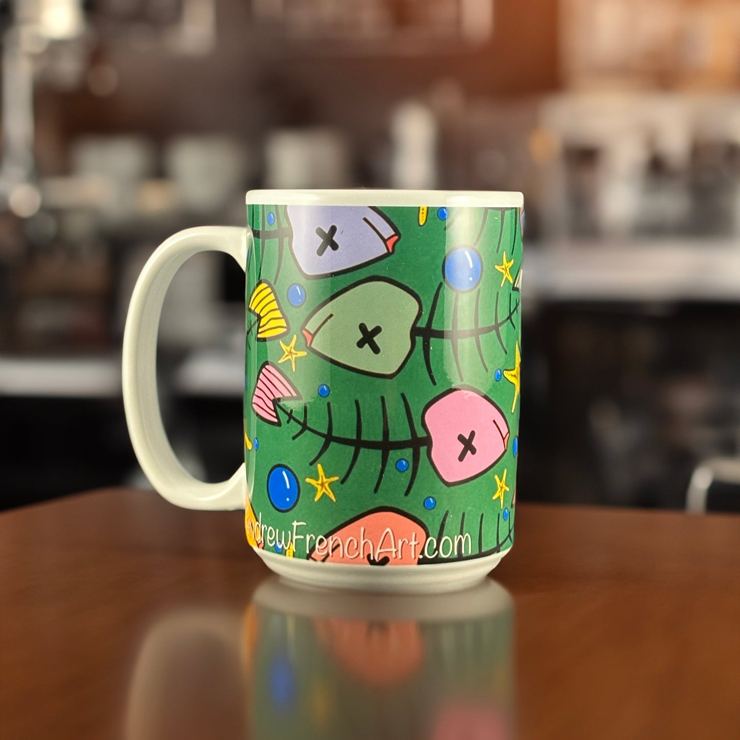 A colorful ceramic mug featuring Andrew French’s “Toxic Xmas Green Fish” design. The artwork showcases whimsical fish bones, seashells, and starfish arranged in a Christmas tree shape on a festive green background — a quirky blend of ocean life and holiday spirit.